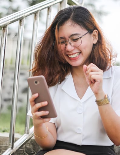 A joyful young woman wearing glasses and a white shirt is happily interacting with her smartphone, which could suggest she's using the Bountisphere personal finance management app, while sitting on outdoor steps with metal railings in the background.