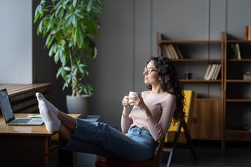 A relaxed woman sits at a desk with her feet up, holding a cup of coffee and gazing out of the window with a calm, confident expression. Her laptop is open on the desk, and the room has a peaceful, modern atmosphere with natural light and a large plant in the background.