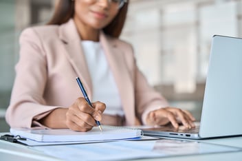 A woman writing in a notebook while using a laptop, representing financial planning and taking control of personal finances.